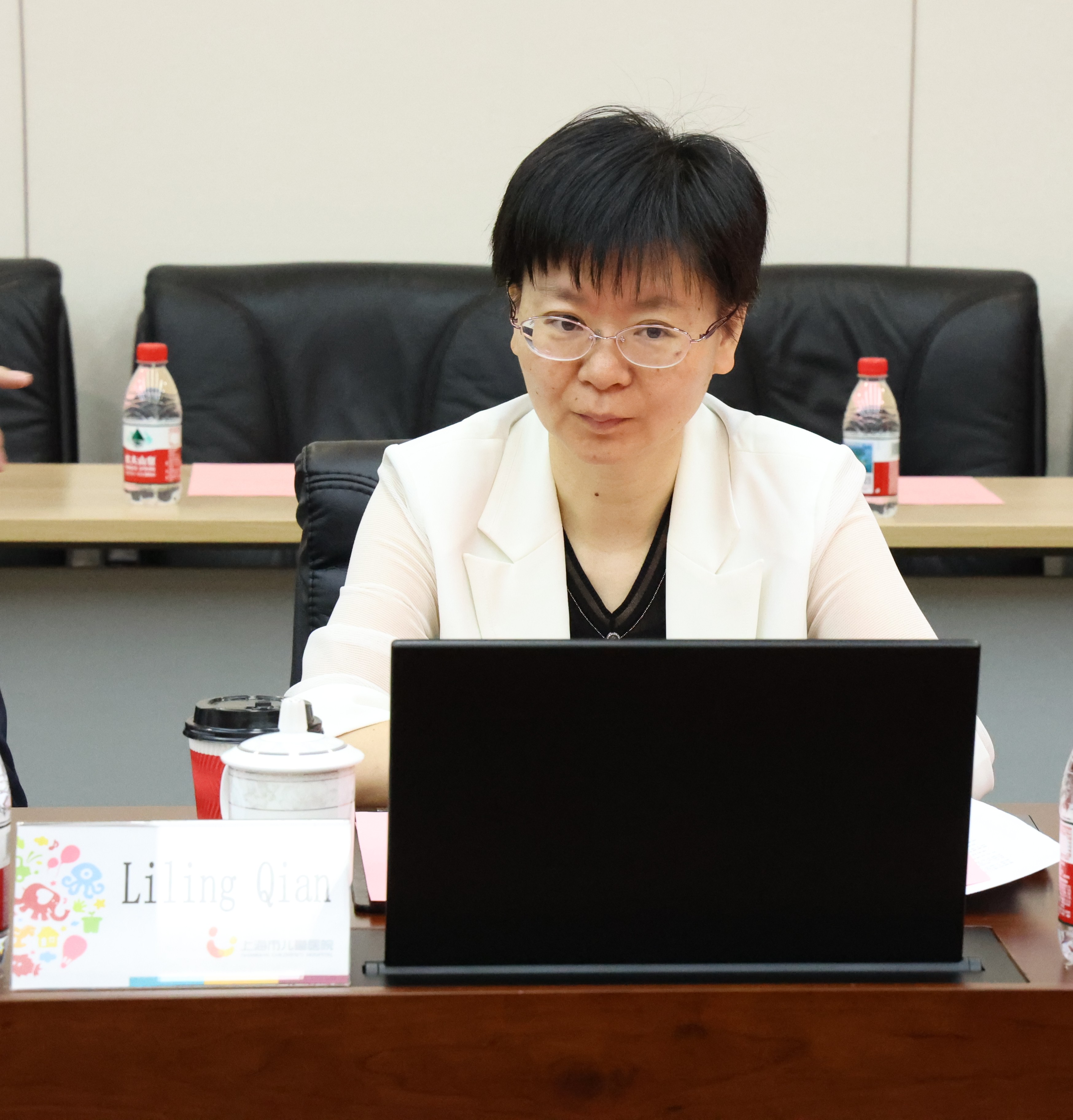 The image shows a person wearing a black suit and a white blouse, sitting at a table with a laptop, surrounded by red and blue chairs, and a water bottle with a lid on the table.AI-generated content may be incorrect.
